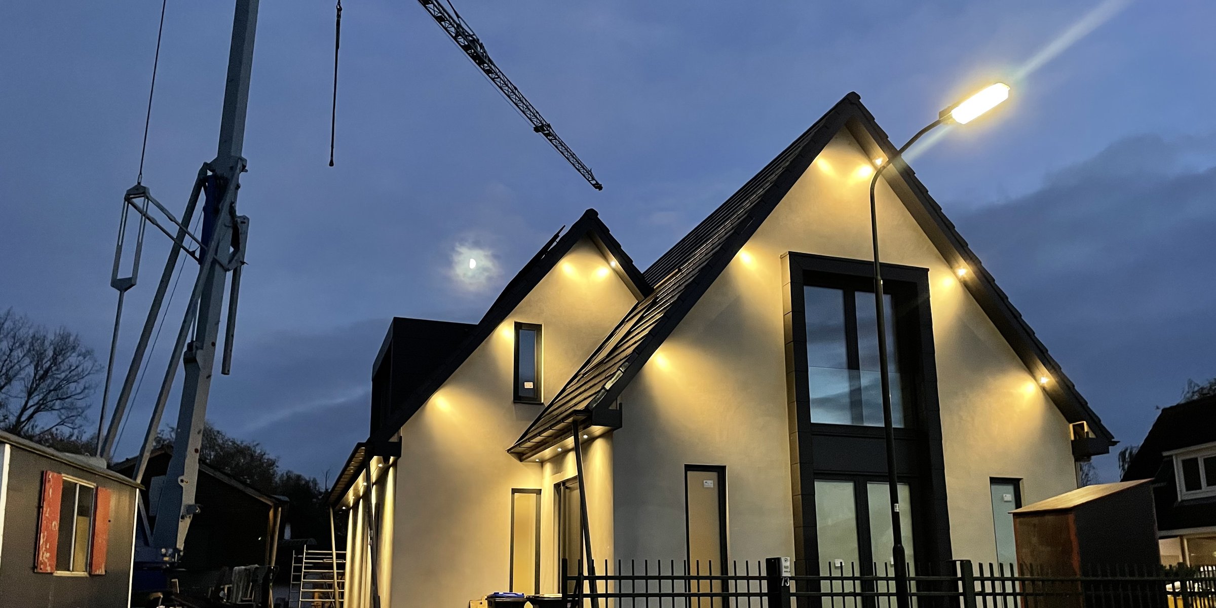 Nieuwbouwwoning in de schemering met brandende gevelspots, bouwkraan op de achtergrond